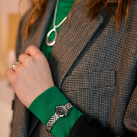 A close-up lifestyle shot of a person wearing a silver mesh-link watch with a round black dial on their wrist, paired with a vibrant green shirt and a gray plaid blazer.