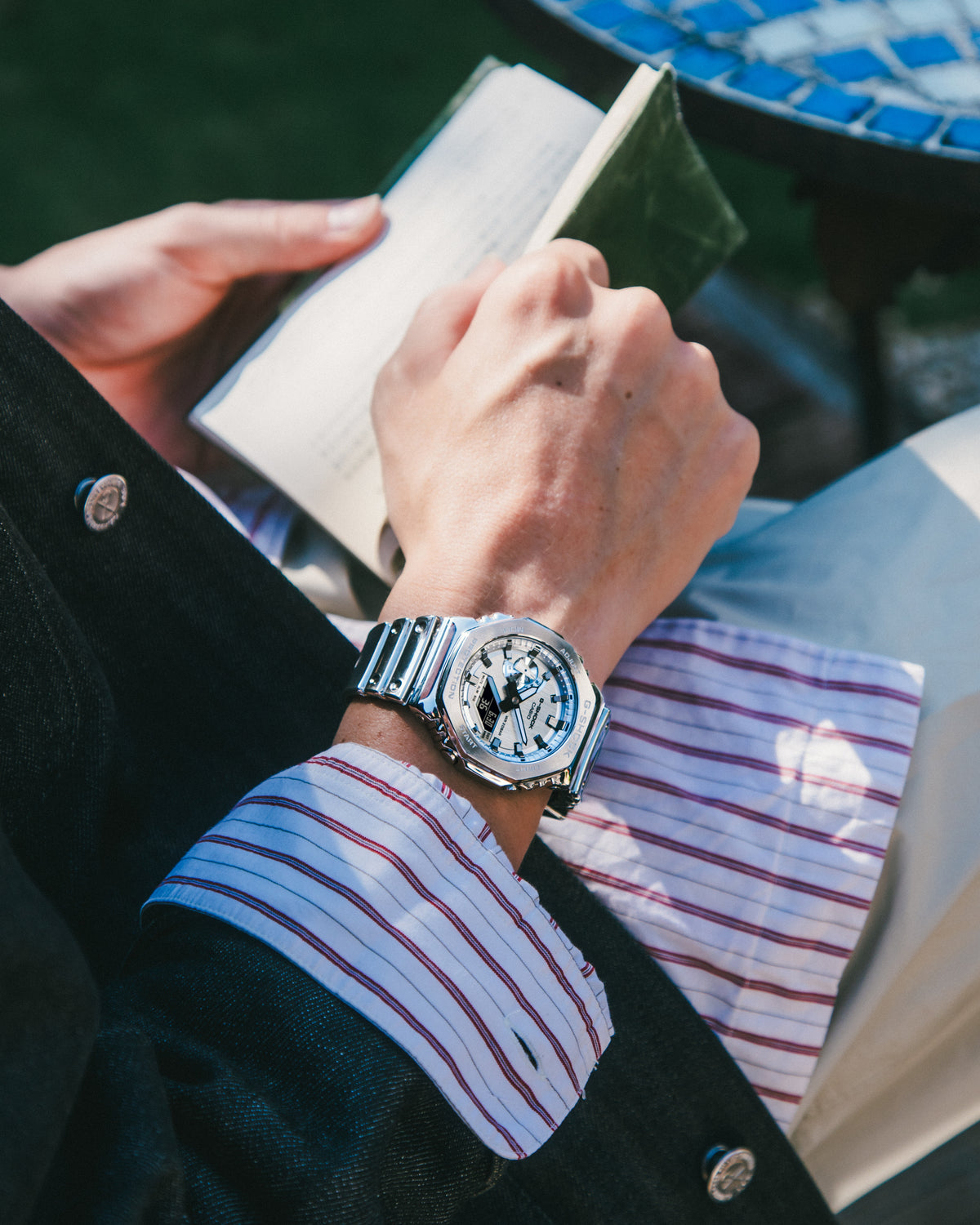 Person wearing a silver watch with a striped shirt sleeve, sitting outdoors.